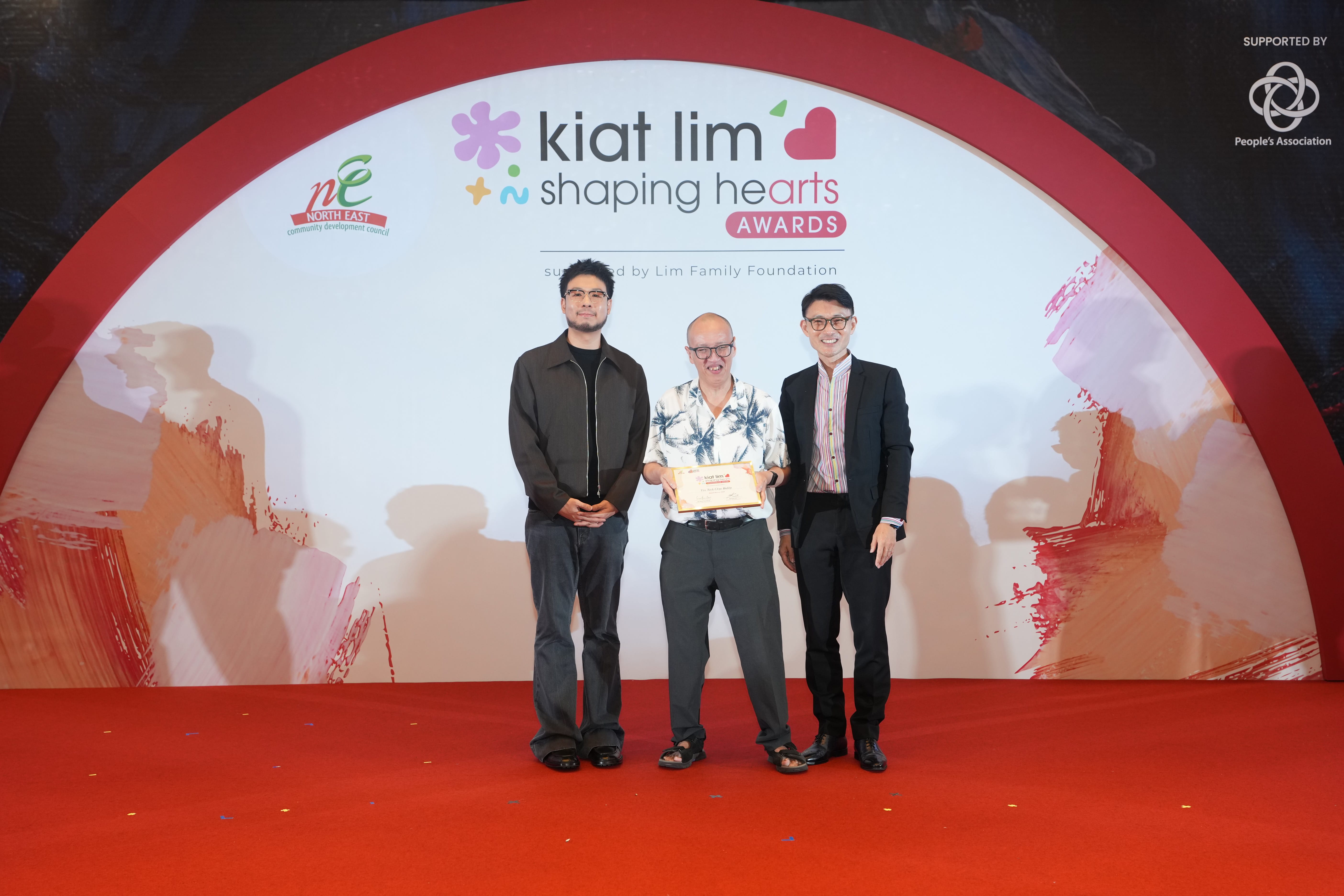 Award recipient standing on stage with the presenter, holding their certificate and posing for a group photo during the award ceremony, with a backdrop reading ‘Kiat Lim-Shaping Hearts Awards'.